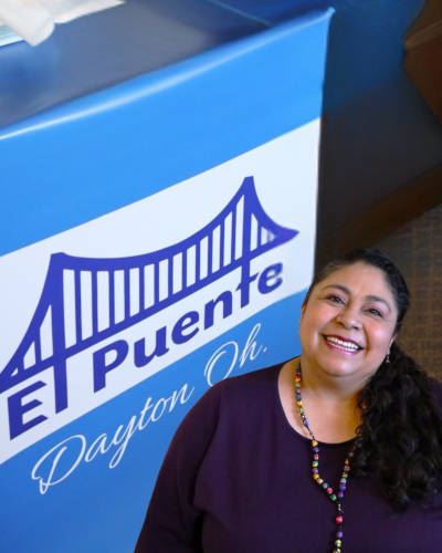 Imelda Ayala Ramos posing for photo by sign with El Puente Educational Center logo, where she works.
