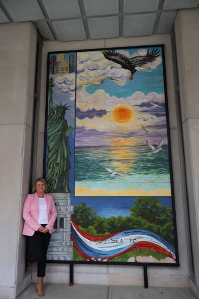 Jeannette Horwitz standing by a mural on a building in downtown Dayton depicting various scenes.