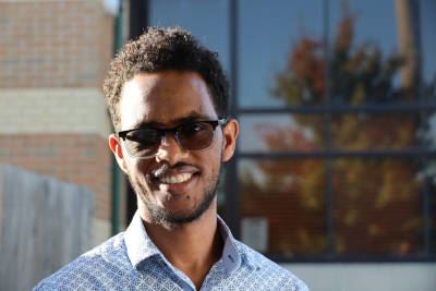 Tesfalem Mahari smiling for photo outdoors in front of building with windows reflecting fall trees.