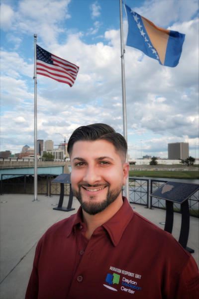 Aldin Fafulovic smiling in front of US and Bosnian flags on poles with Dayton in the background.
