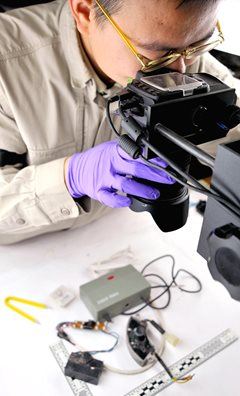 Lab assistant wearing purple gloves examines electrical components under a camera/microscope.